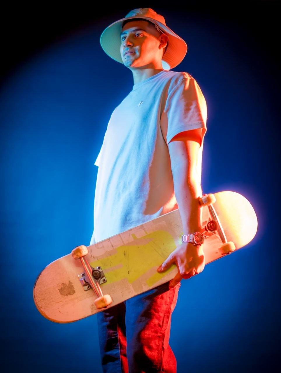 Kevin Lucano holding a skateboard, wearing a hat, against a blue-lit backdrop.